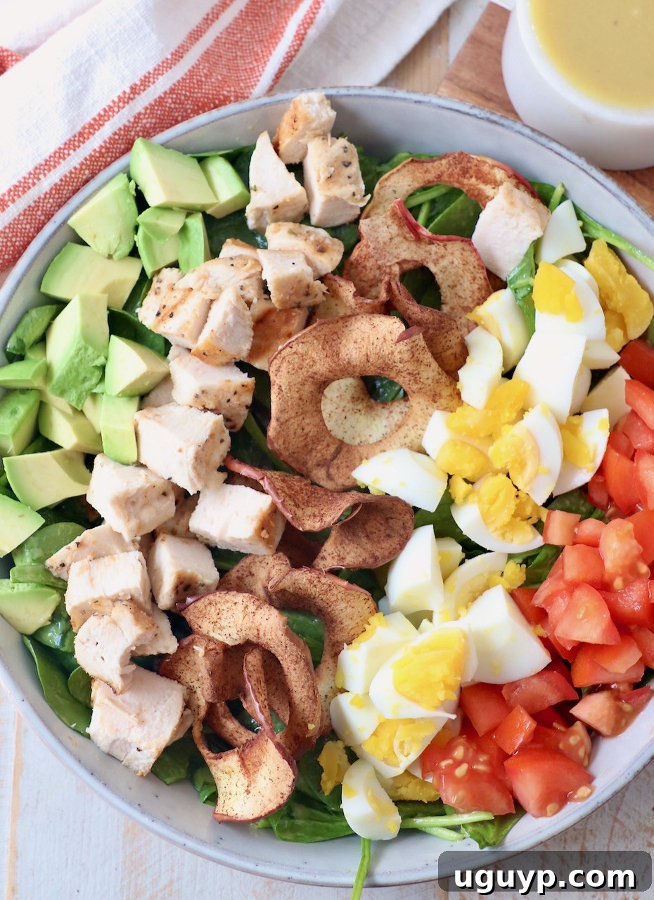 A vibrant, healthy Cobb Salad in a bowl, artfully topped with golden apple chips, perfectly cooked hard-boiled eggs, juicy tomatoes, tender grilled chicken, and creamy avocado slices. The diverse textures and colors create an inviting and wholesome meal.
