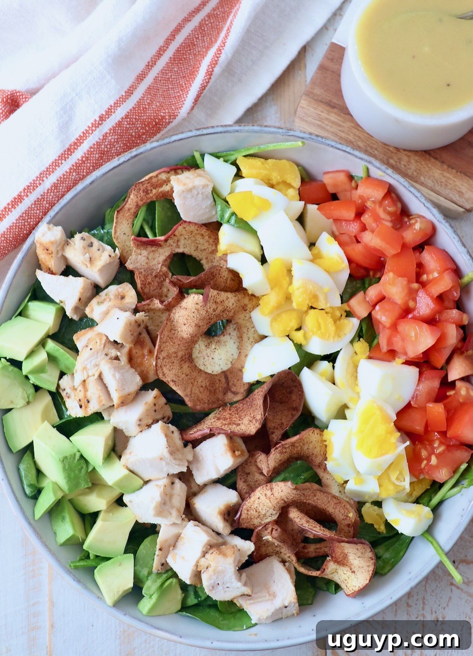 A beautifully assembled Cobb Salad, clearly showing layers of spinach, grilled chicken, hard-boiled eggs, tomatoes, avocado, and apple chips, with a small bowl of homemade honey dijon dressing on the side, ready to be drizzled.