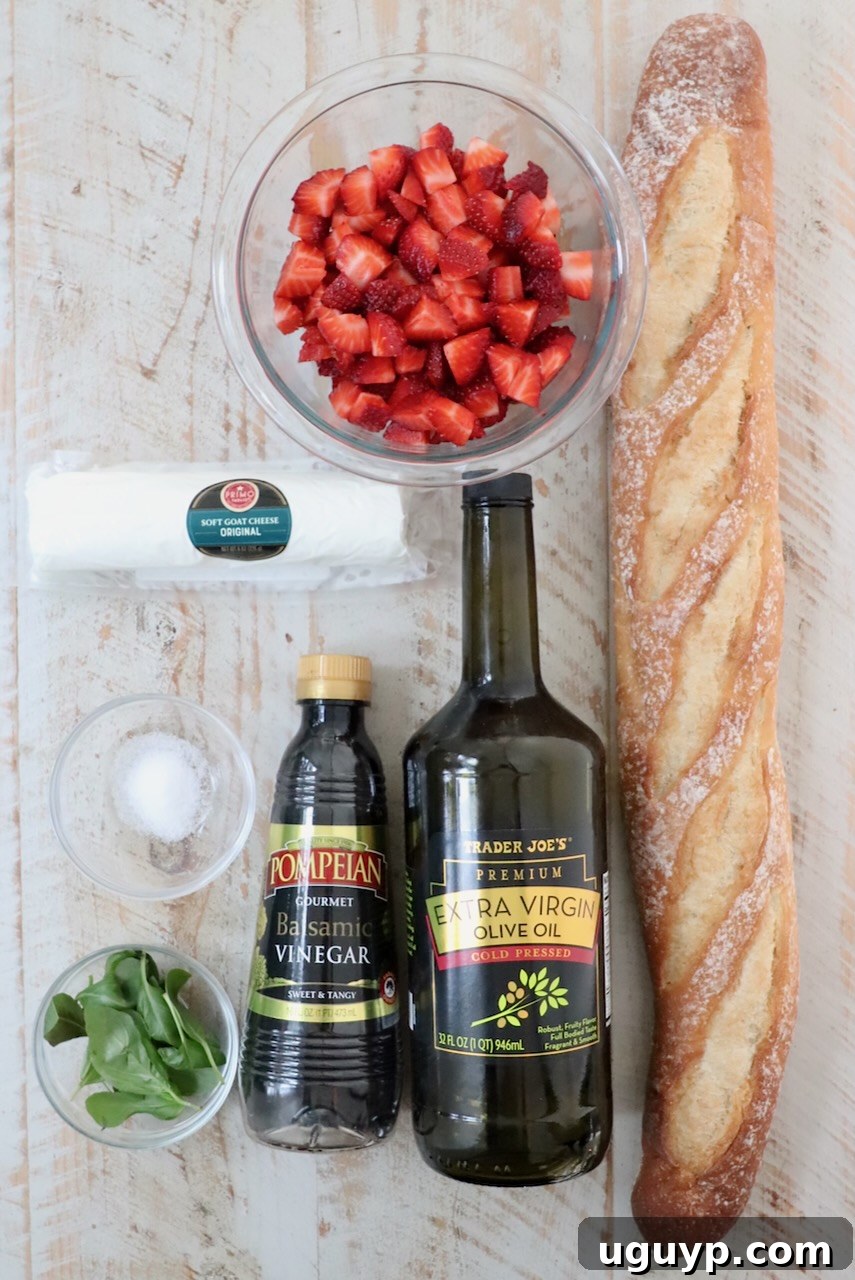 All the fresh ingredients for strawberry balsamic bruschetta, including strawberries, basil, balsamic vinegar, goat cheese, and baguette, artfully arranged on a white wood board.