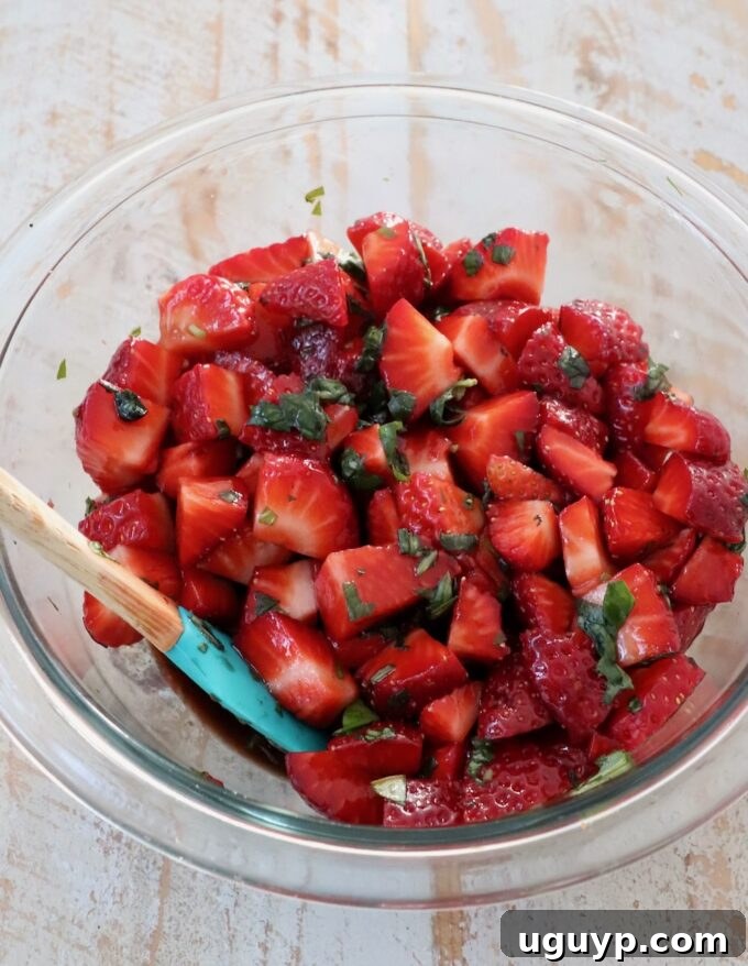 Freshly diced strawberries in a clear mixing bowl, with balsamic vinegar and vibrant fresh basil leaves, ready to be combined.