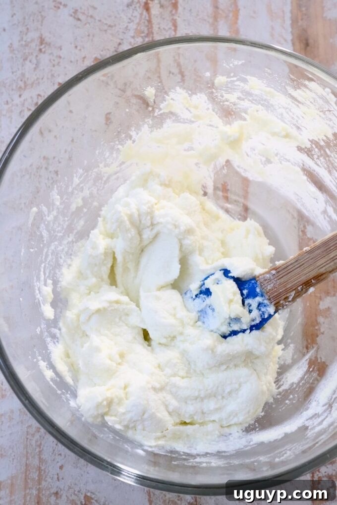 Creamy goat cheese being whipped in a glass bowl with a blue spatula, achieving a smooth and spreadable consistency.