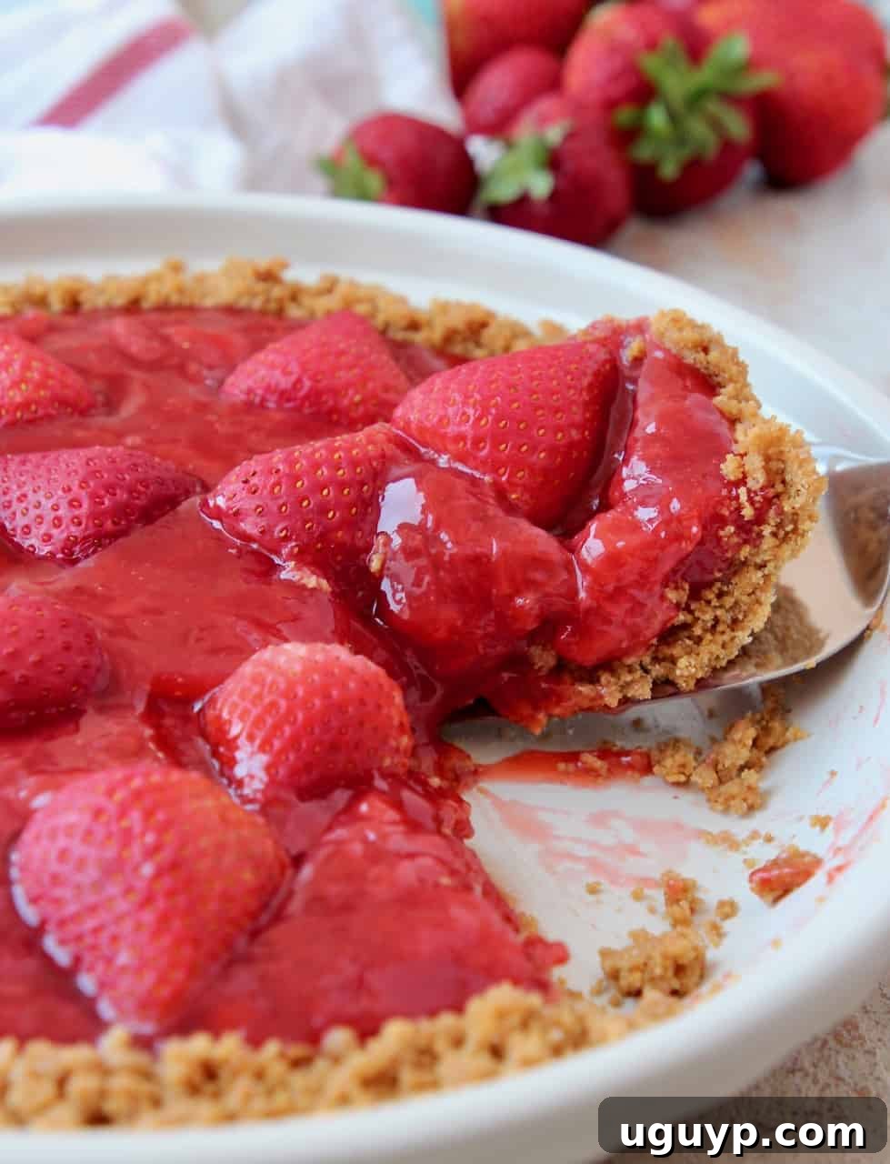 A perfectly sliced piece of fresh strawberry pie being lifted from a pie plate, showcasing the vibrant red filling and golden graham cracker crust.