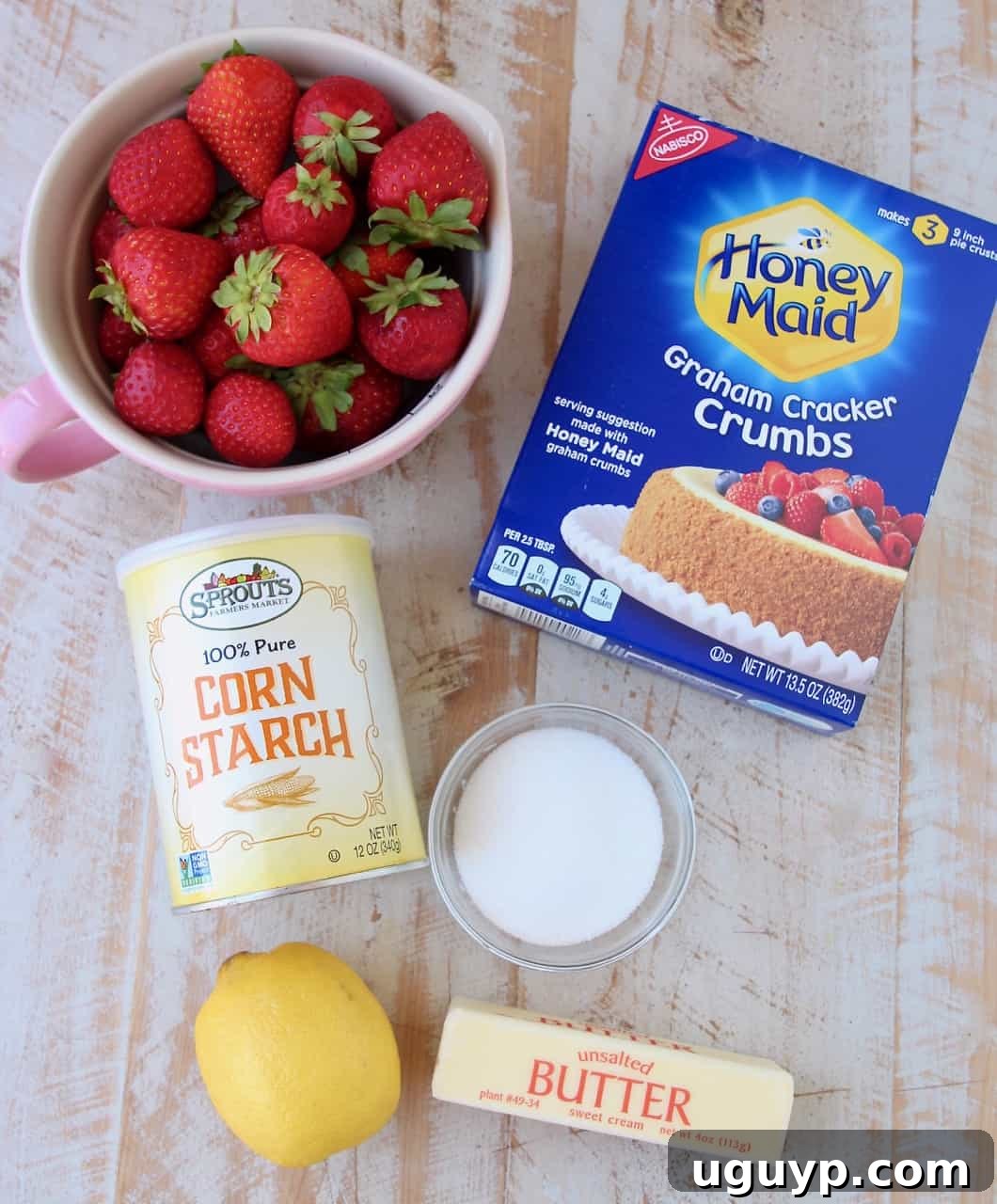 Fresh strawberry pie ingredients laid out on a kitchen counter, including whole strawberries, sugar, cornstarch, and graham cracker crumbs.