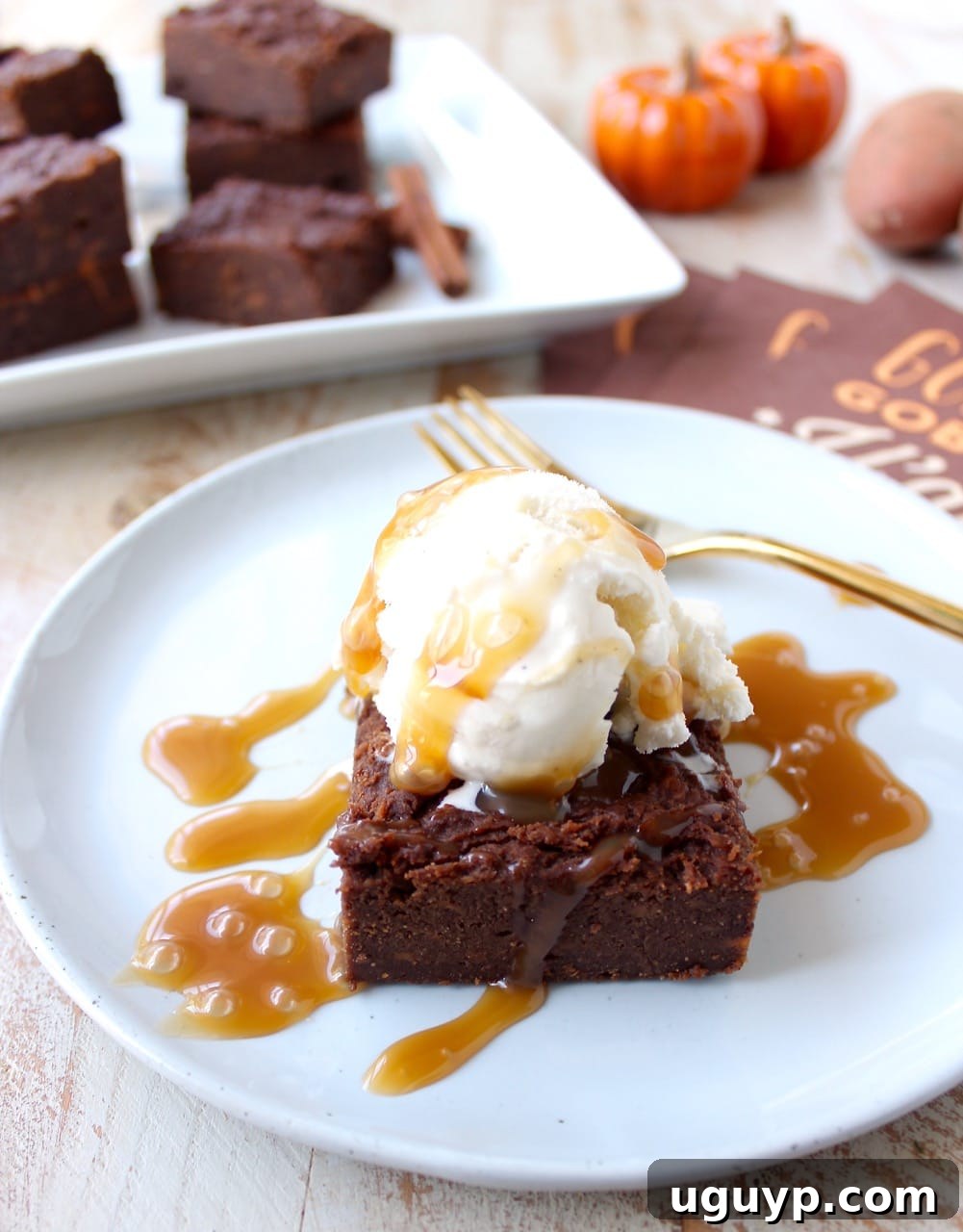 A beautifully presented slice of gluten-free sweet potato brownie, topped with vanilla ice cream and caramel, on a white plate.