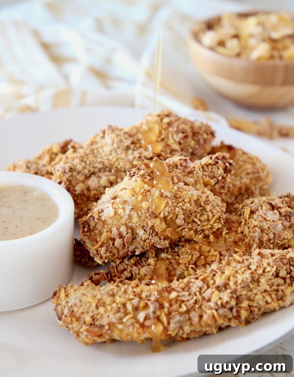 Baked honey bunches of oats chicken strips piled on plate with honey drizzle over the chicken and bowl of honey bunches of oats cereal in background