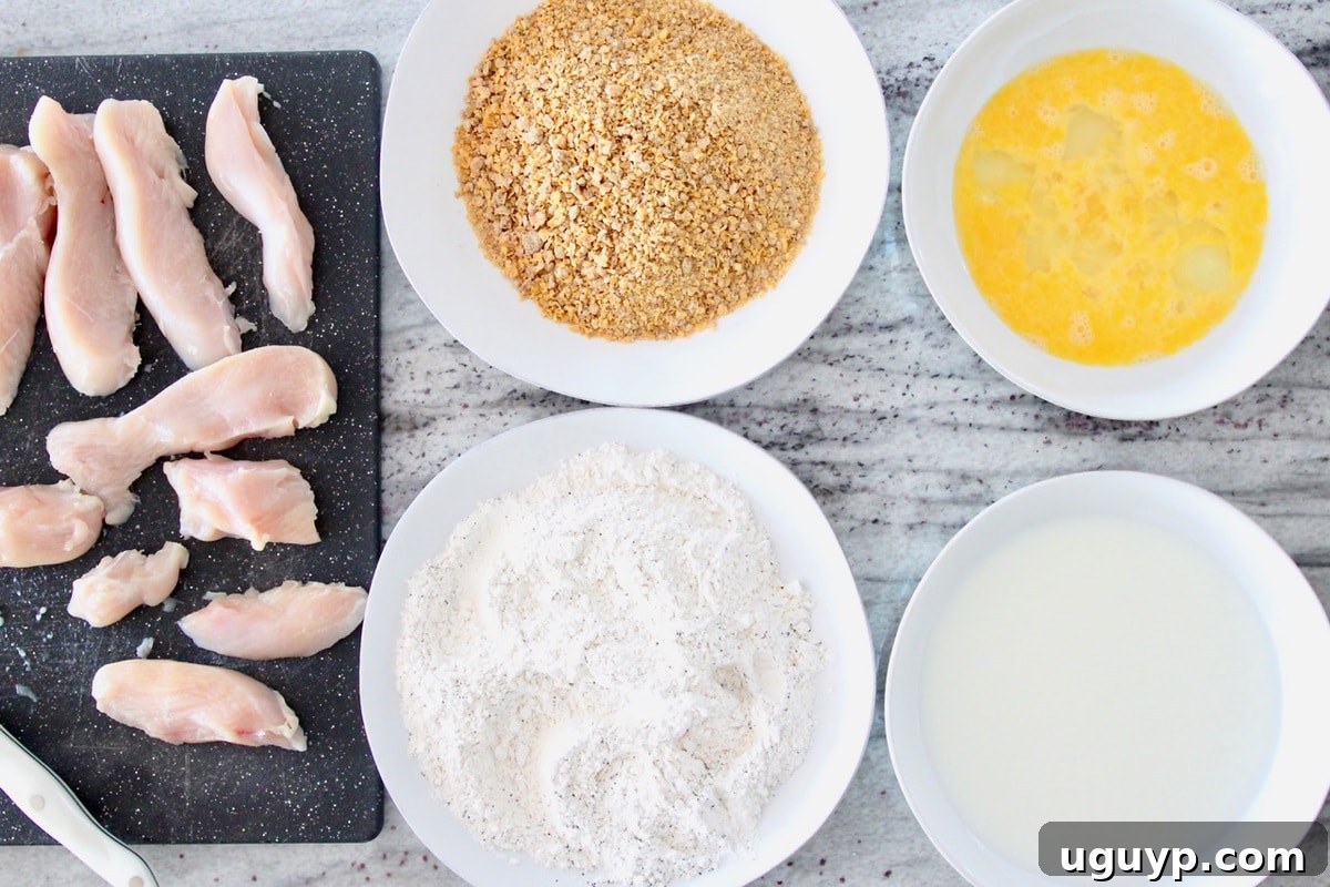 Chicken strip batter station with bowl of eggs, bowl of milk, plate of flour, plate of crushed cereal and raw chicken on cutting board