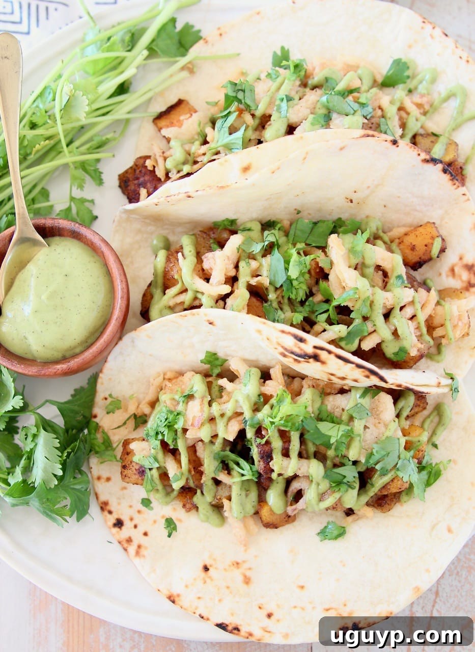 Butternut squash tacos in flour tortillas on plate with small bowl of creamy avocado sauce and fresh cilantro