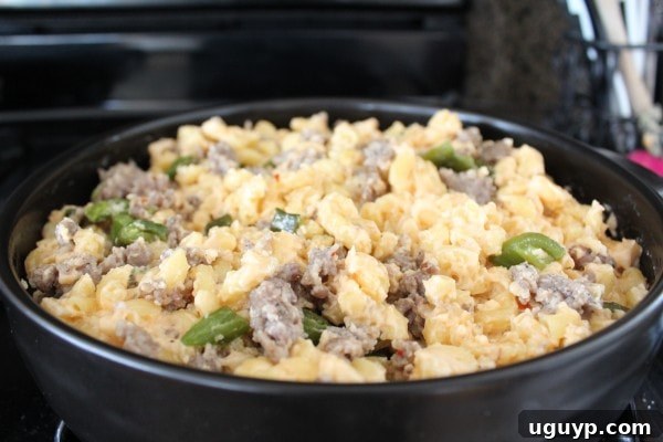 Diced roasted jalapeños and browned sausage being mixed into the creamy macaroni and cheese in a skillet, combining all the flavors.