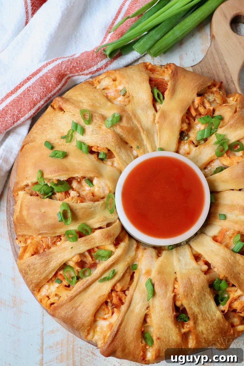 Golden brown buffalo chicken crescent roll ring on a rustic wooden serving tray with a small bowl of buffalo sauce in the center.