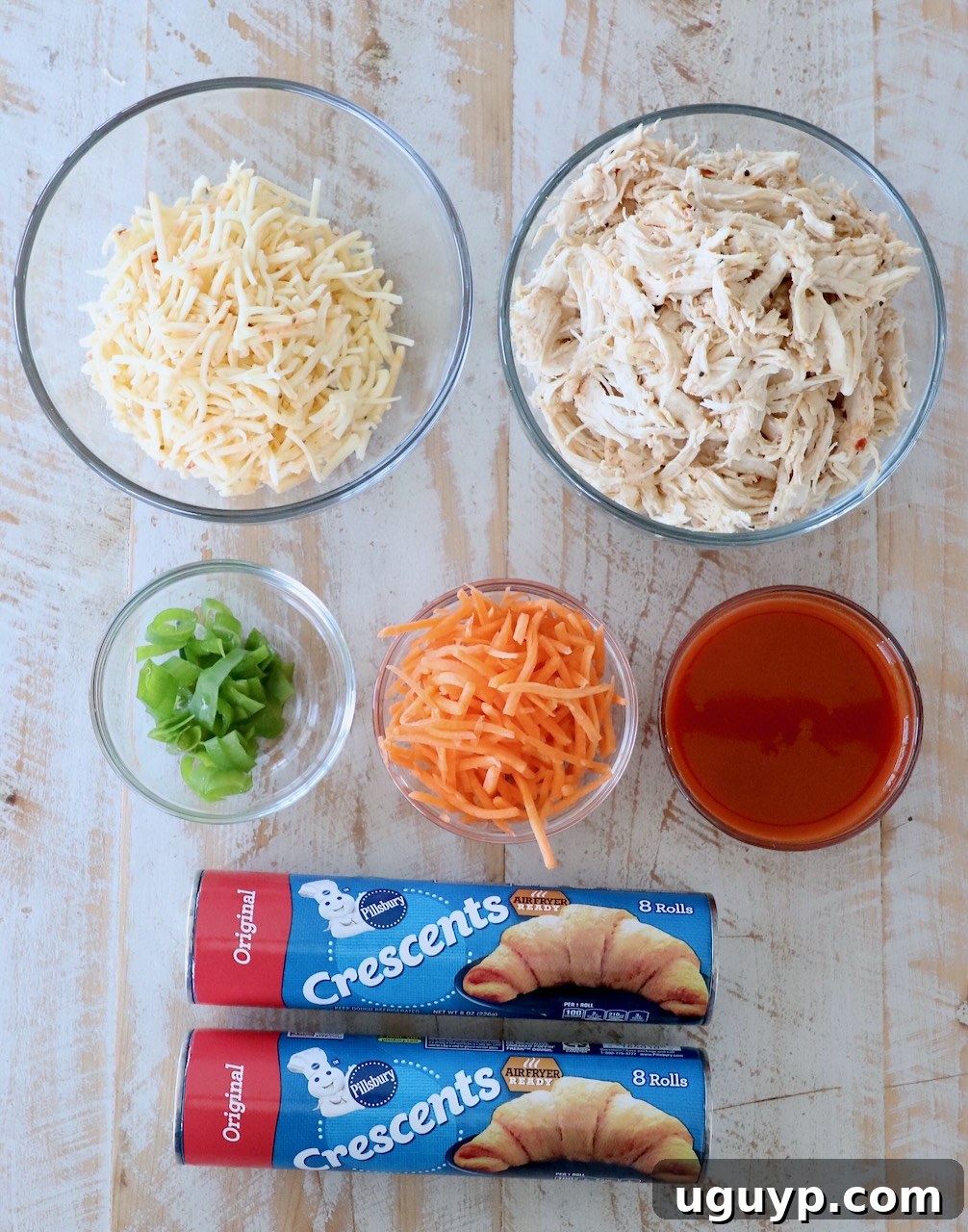 Arrangement of ingredients for buffalo chicken crescent ring on a white wooden cutting board, including shredded chicken, buffalo sauce, cheese, carrots, and crescent roll dough.