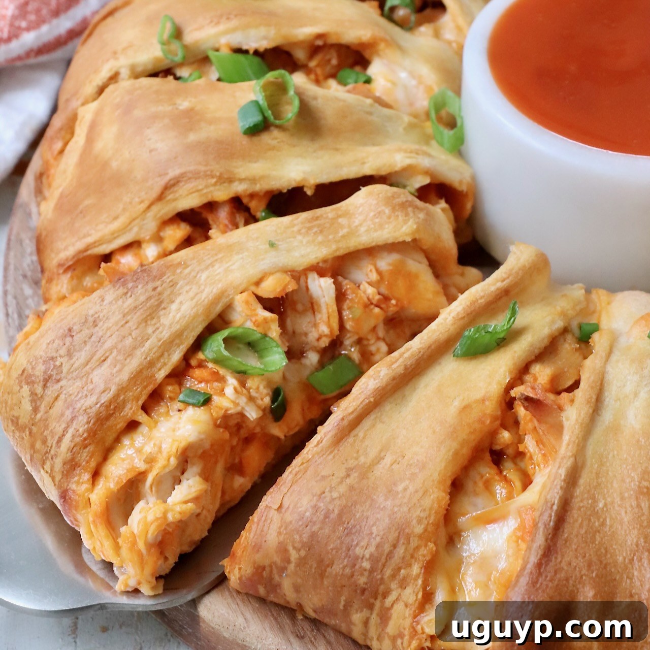 A slice of buffalo chicken crescent ring on a spatula, with a small bowl of buffalo sauce and a sprinkle of green onions beside it.