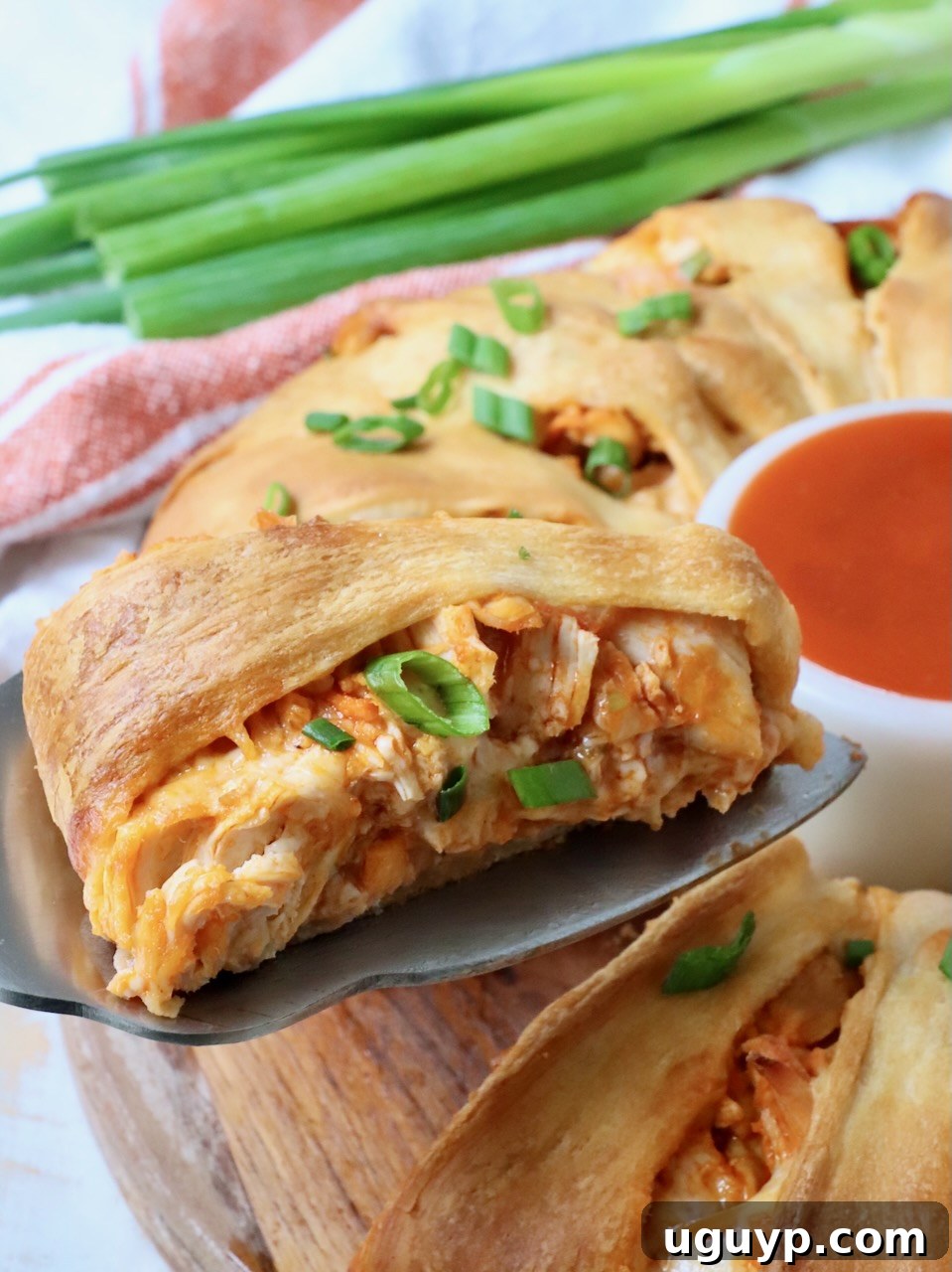 Close-up of a delicious slice of buffalo chicken crescent ring on a spatula, garnished with fresh green onions, with a small bowl of buffalo sauce in the background.