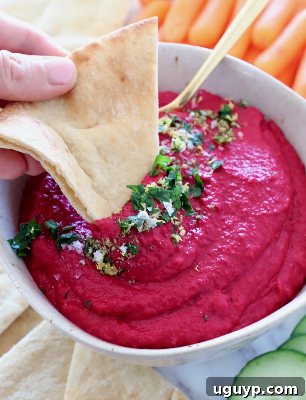 A bowl of vibrant beet hummus beautifully garnished, with a piece of pita bread dipped in.