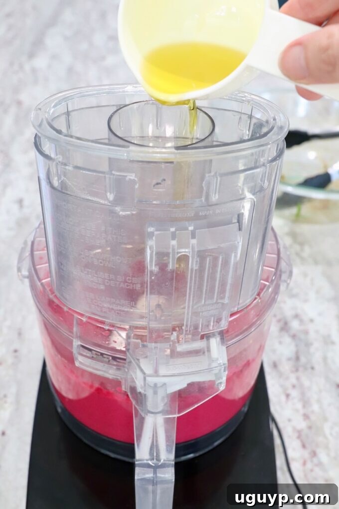 Olive oil being slowly drizzled into a food processor while blending beet hummus.