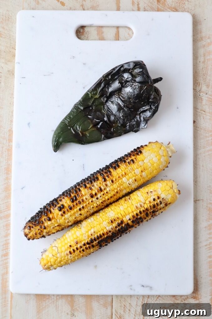 Grilled corn on the cob and a roasted poblano pepper resting on a wooden cutting board.