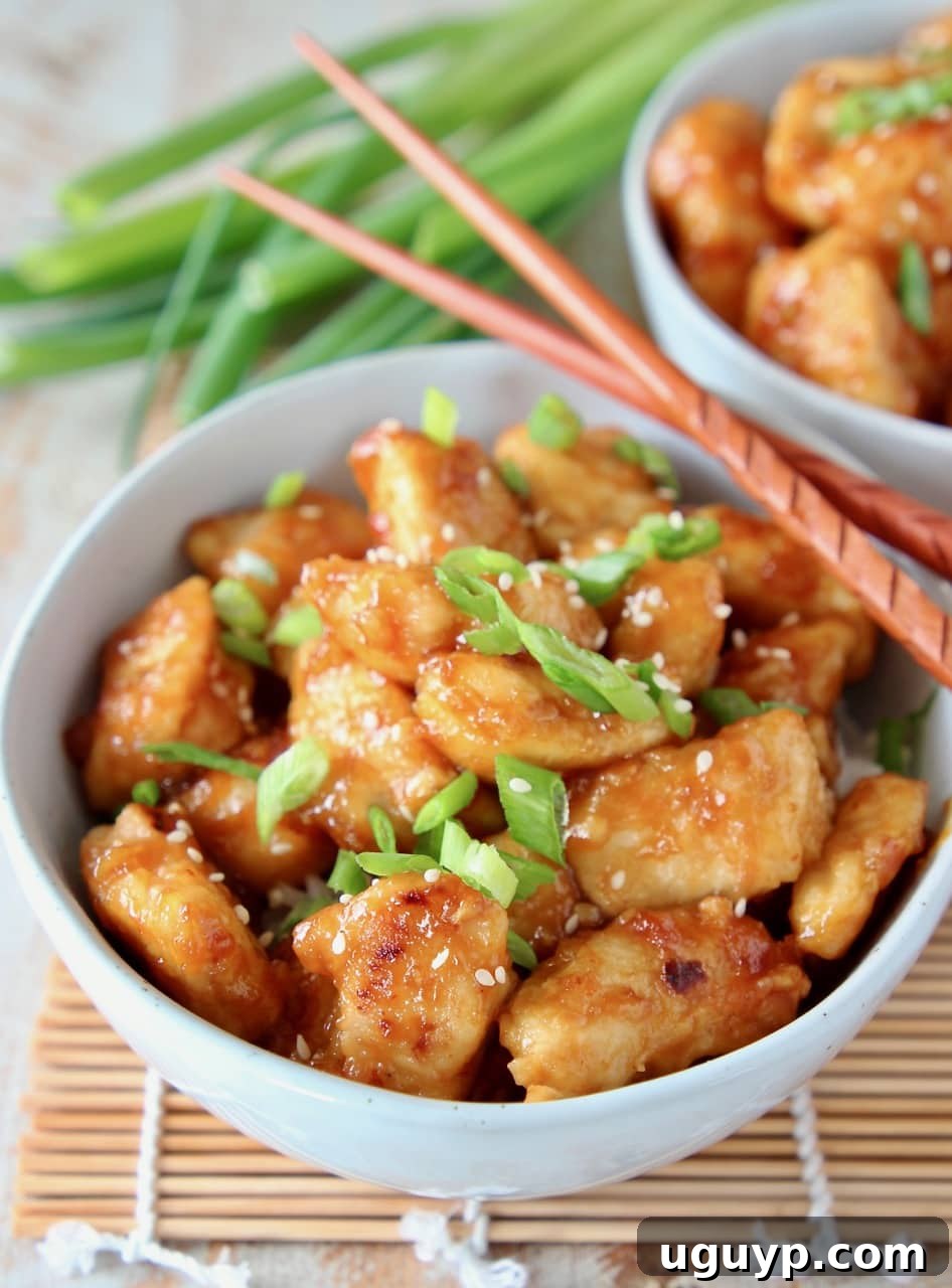 Orange chicken in bowl topped with sliced scallions and sesame seeds, with wood chopsticks on the side of the bowl
