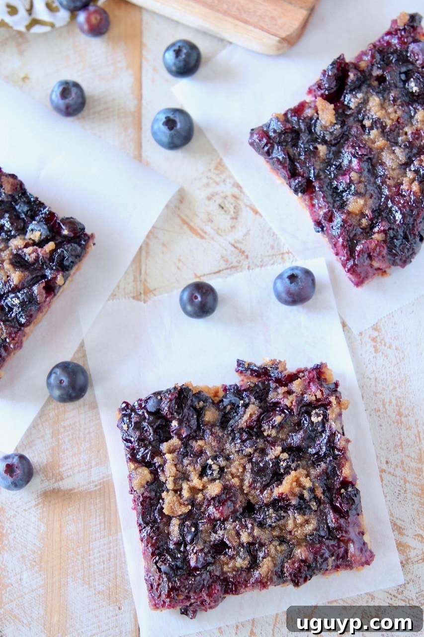 A vibrant overhead shot of freshly baked blueberry oat bars, cut into neat squares and arranged on white parchment paper, adorned with scattered fresh blueberries.
