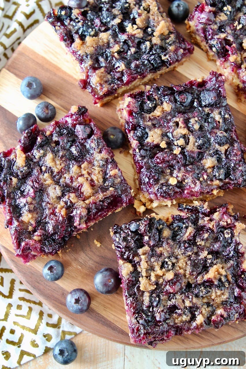Individual gluten-free blueberry oat bars, perfectly cut into squares, arranged on a wooden cutting board with a scattering of fresh blueberries.
