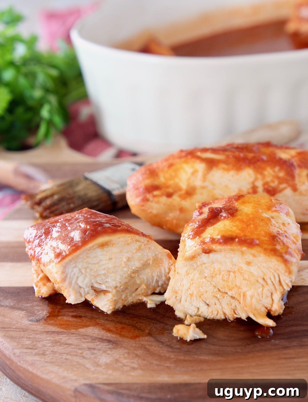 Two succulent pieces of baked chicken on a rustic wood cutting board, one expertly sliced in half to reveal its juicy interior, with a pastry brush in the soft-focus background, ready for basting.