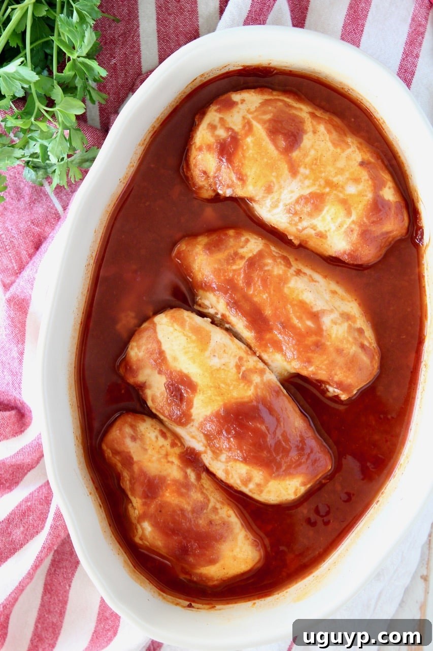 Overhead shot of perfectly baked chicken breasts swimming in a rich bbq sauce, nestled in an oval baking dish, all atop a charming red and white striped towel with fresh parsley elegantly arranged on the side.