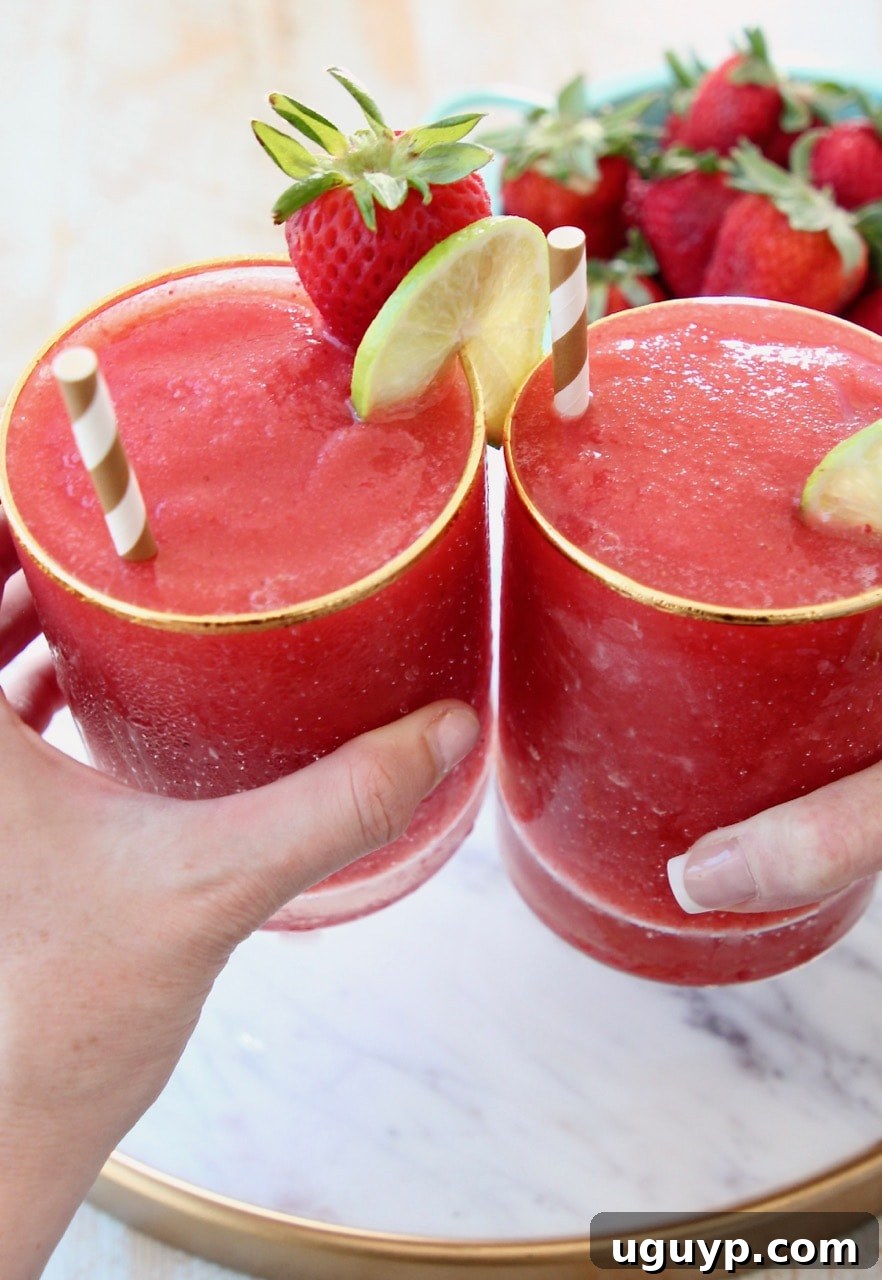 Two hands holding glasses of strawberry daiquiris with gold and white straws and lime wedges on the side