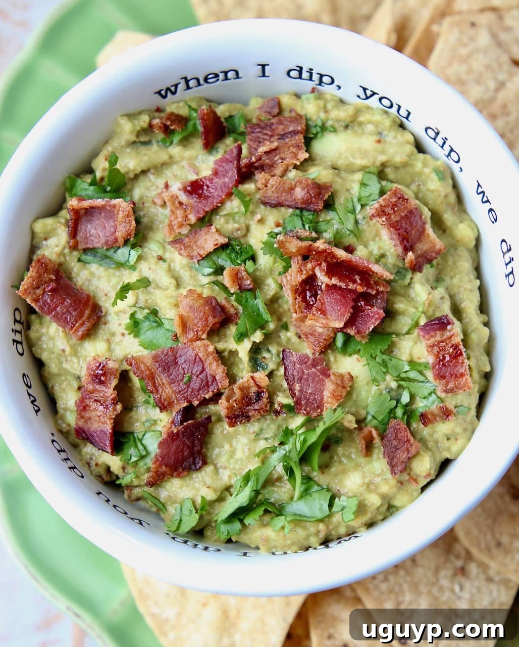 Guacamole in white bowl with crumbled cooked bacon on top, surrounded by fresh ingredients like cilantro and lime.