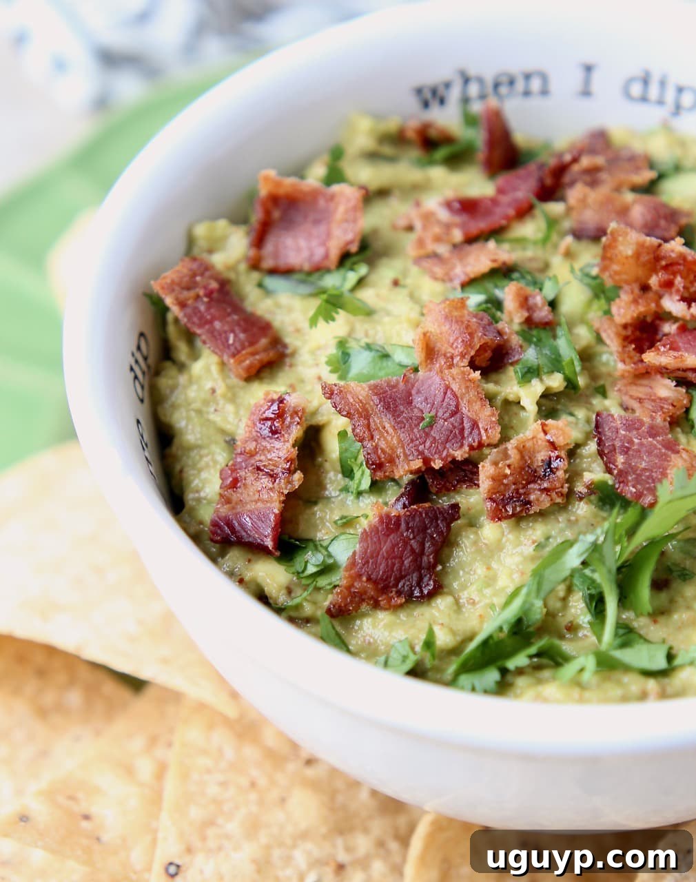 Close-up of creamy Chipotle Bacon Guacamole in a white bowl, garnished with fresh cilantro and crispy bacon pieces.