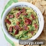 Guacamole in white bowl topped with cooked bacon, with tortilla chips on the side, ready to be enjoyed.