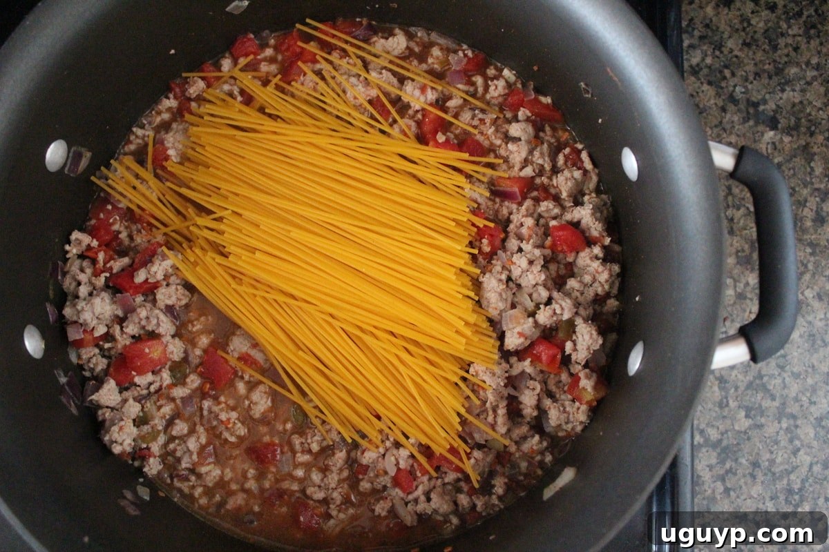 Broken gluten-free spaghetti noodles being added to a simmering pot filled with a savory taco-flavored meat and tomato sauce mixture.