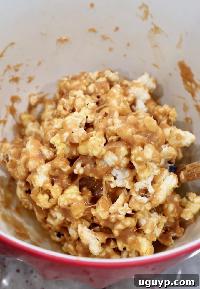The rich peanut butter sauce being tossed with popped popcorn in a large mixing bowl, coating every kernel.