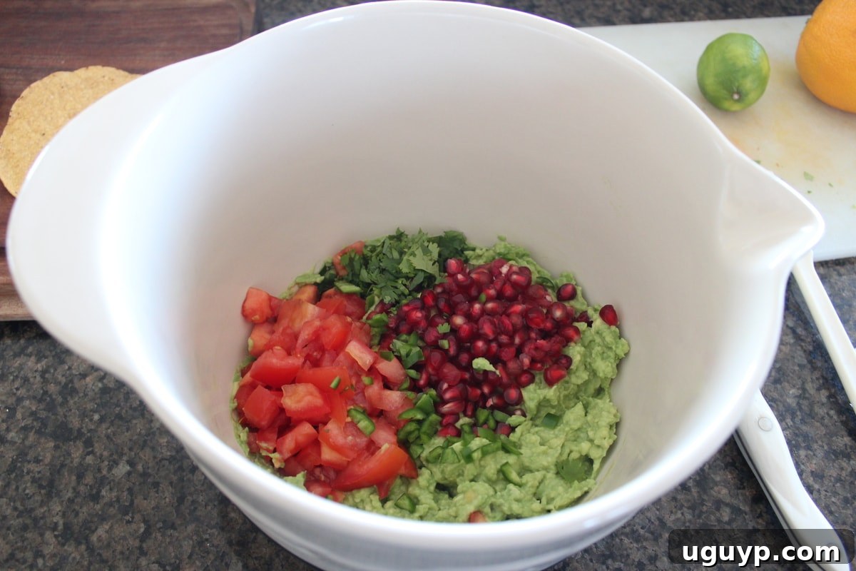 Mashed avocado in a bowl with diced tomatoes and bright red pomegranate arils added, ready to be mixed.