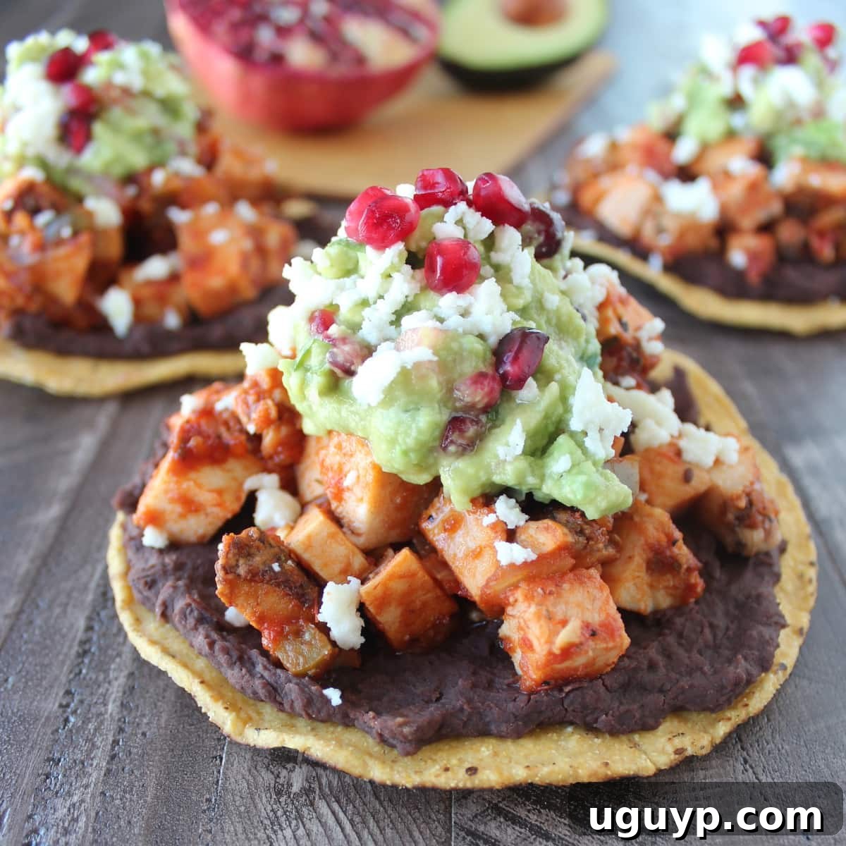 A beautifully assembled turkey tostada with layers of beans, seasoned turkey, and pomegranate guacamole, garnished with queso fresco, on a wooden surface.