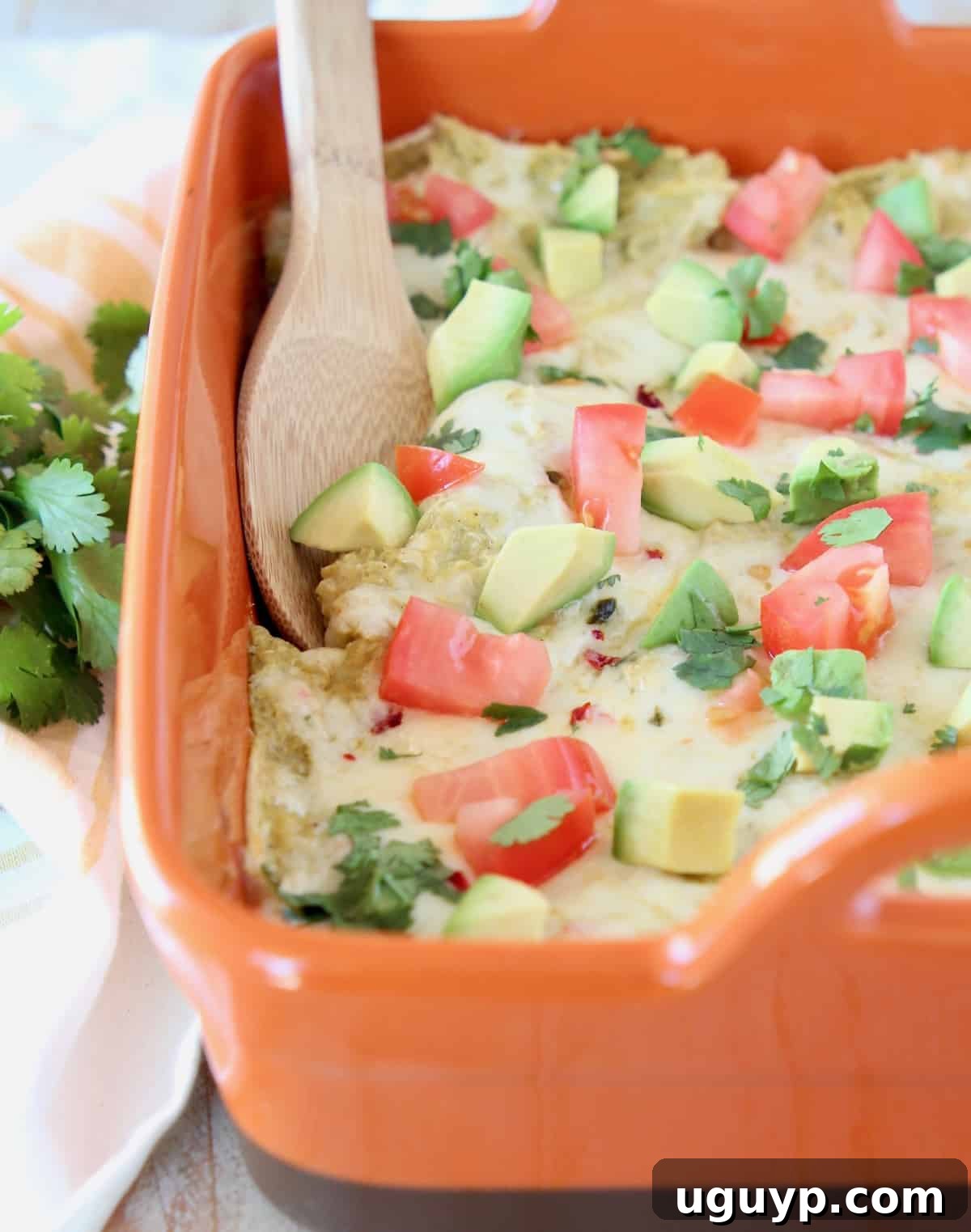 Chicken Enchilada Casserole in an orange baking dish with a wooden spoon, garnished with fresh cilantro and lime wedges