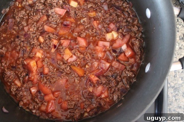 After browning the beef and onions, diced tomatoes and tomato sauce are added to the pot, simmering to create a rich, flavorful base.
