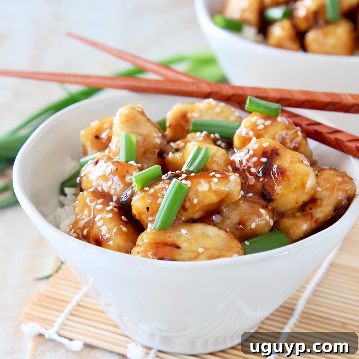 Tender pieces of golden Chinese Honey Chicken nestled in a white bowl with fluffy white rice, adorned with fresh chopped green onions and a sprinkle of sesame seeds. Brown chopsticks rest elegantly beside the bowl, inviting you to dig in.