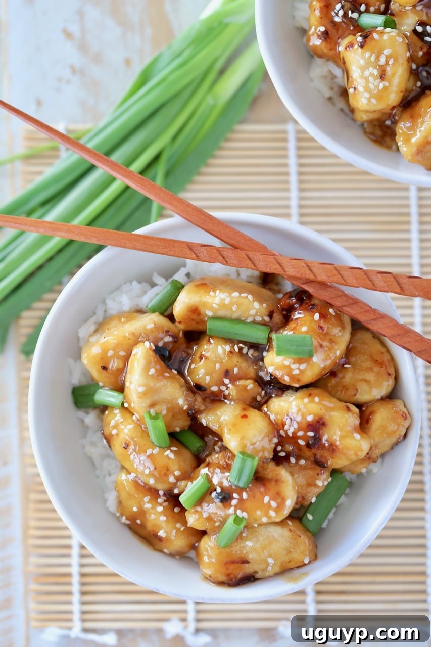 An inviting overhead shot of Chinese honey chicken presented in a white bowl, accompanied by a portion of white rice. The dish is beautifully garnished with fresh green onions and sesame seeds, with elegant brown chopsticks placed on the side. Additional green onions are artfully arranged next to the bowl in the background.