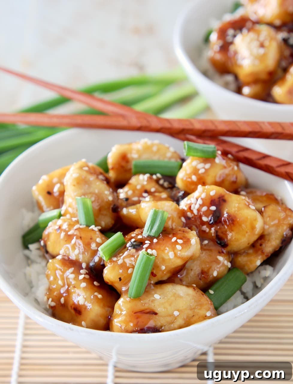A bowl of Chinese honey chicken, garnished with green onions and sesame seeds, sits next to a serving of white rice. Brown chopsticks are placed on the side of the bowl, with whole green onions blurred in the background, creating an appetizing scene.