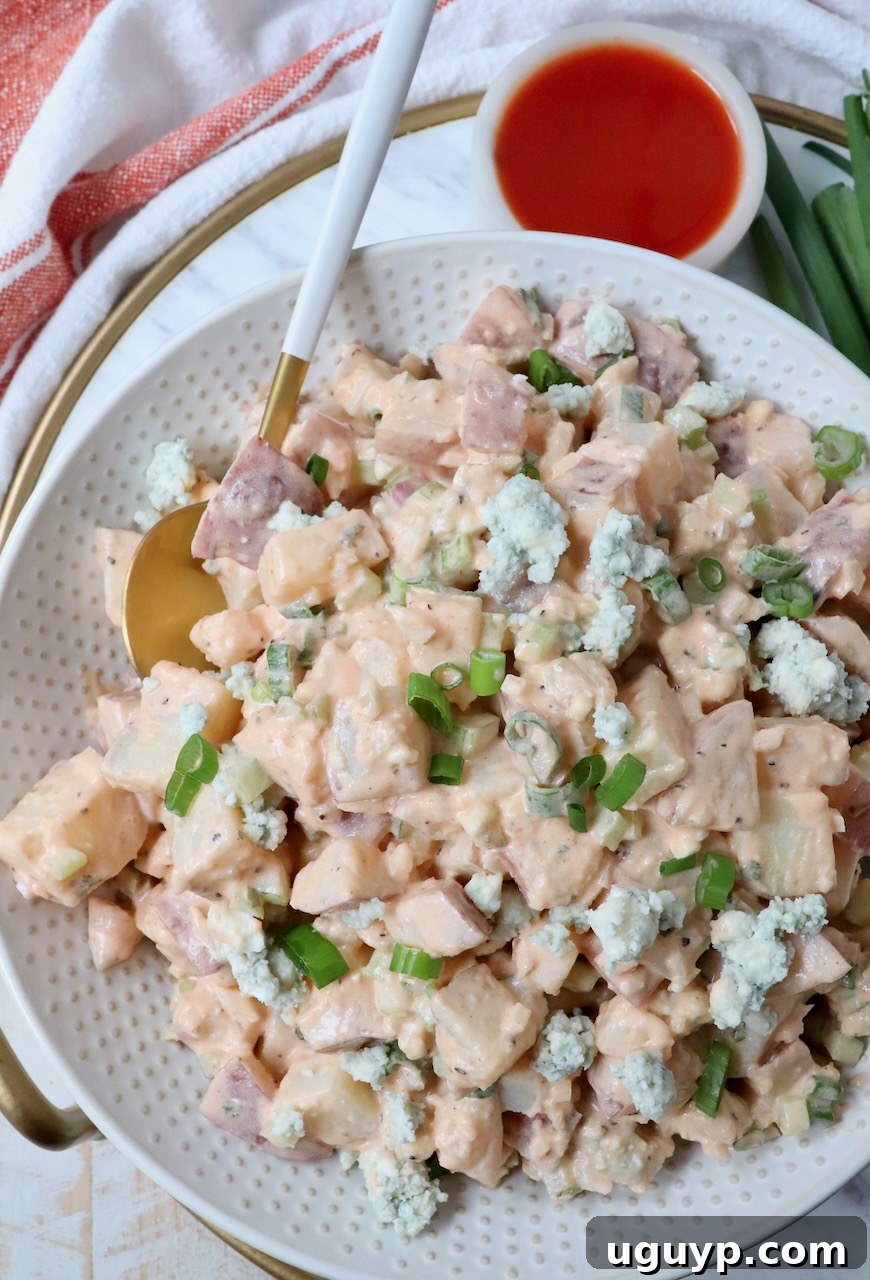 Close-up of Buffalo Potato Salad in a white bowl, with a spoon and a separate small bowl of buffalo sauce on a rustic wooden surface, highlighting the creamy texture.