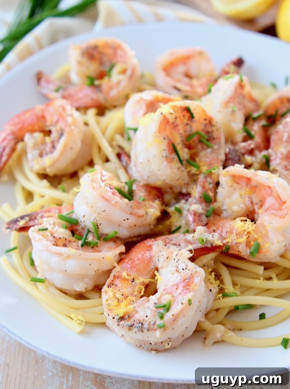 Close-up shot of cooked shrimp scampi over pasta, elegantly topped with fresh chives and lemon zest, served on a white plate with a fork.