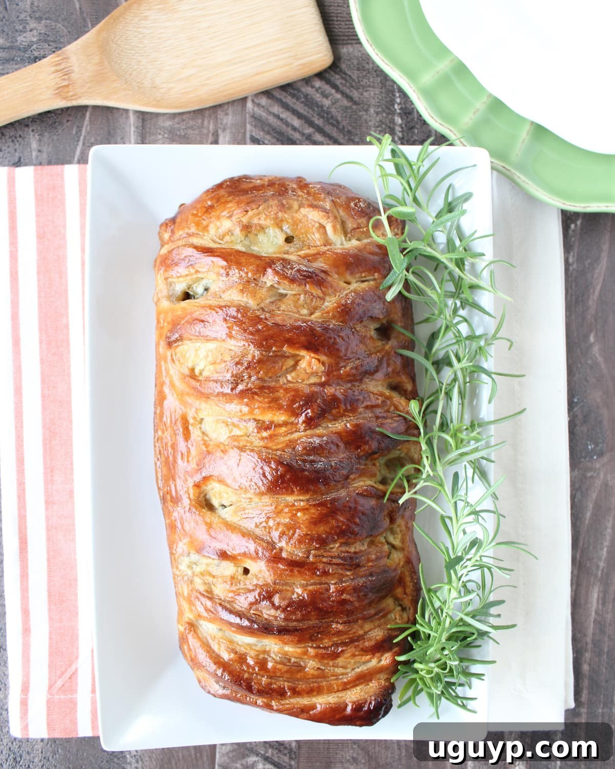 A freshly baked, braided, and filled puff pastry on a plate, garnished with fresh rosemary, steam gently rising.