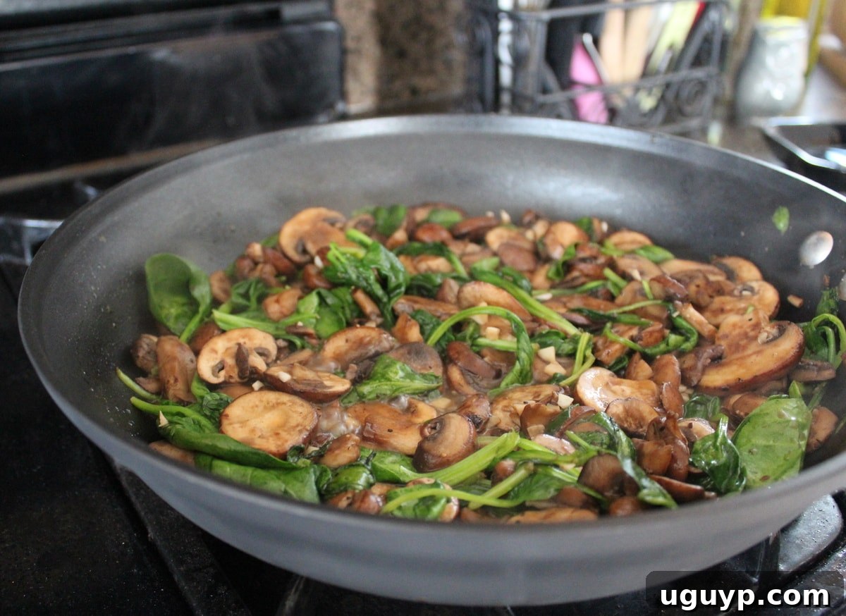 Sliced mushrooms and fresh spinach leaves cooking in a large skillet on the stove, seasoned with salt and pepper.