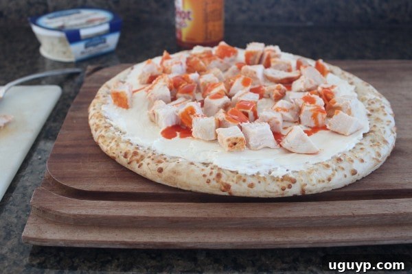 A pizza crust topped with a white cheese sauce, diced buffalo chicken, and drizzled with more buffalo sauce on a wooden cutting board.