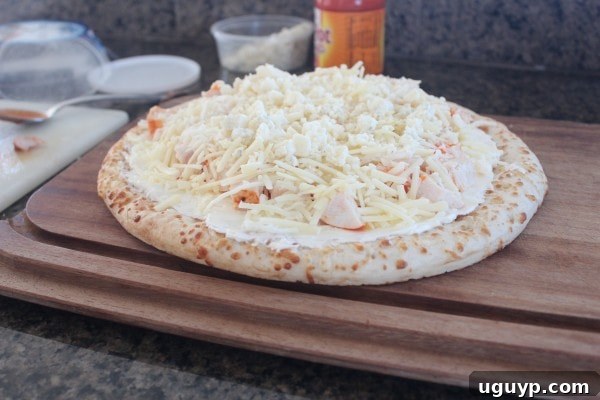 An uncooked Buffalo Chicken Dip Pizza, generously topped with three types of cheese, on a wooden cutting board.