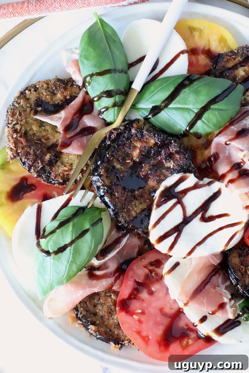 A vibrant Caprese salad with sliced tomatoes, mozzarella, crispy eggplant, prosciutto, fresh basil, and balsamic glaze drizzle on a white plate.
