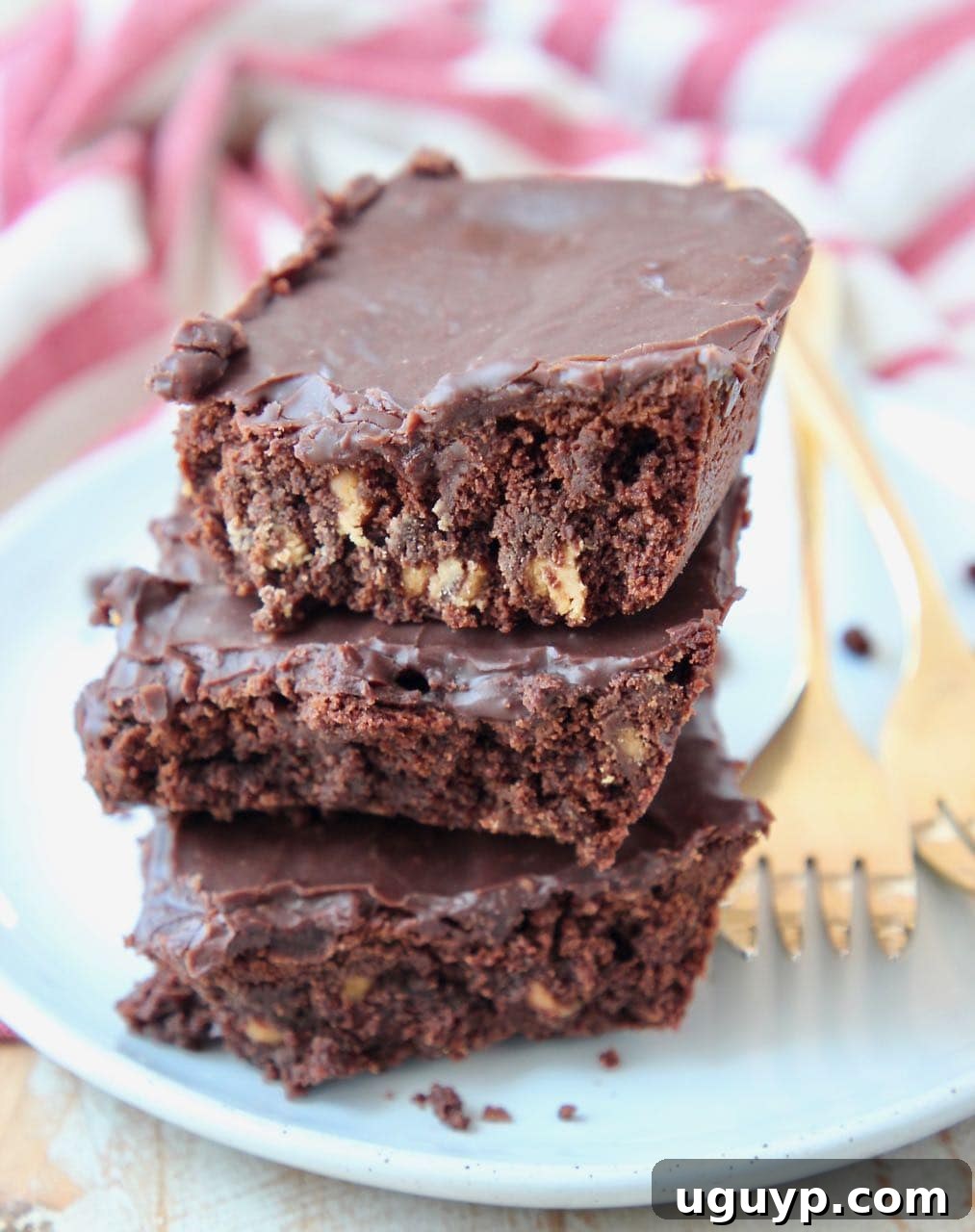Three stacked brownies on a white plate with gold forks on the side