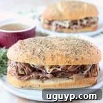 Tri tip sandwich on plate with cup of au jus and fresh rosemary garnish