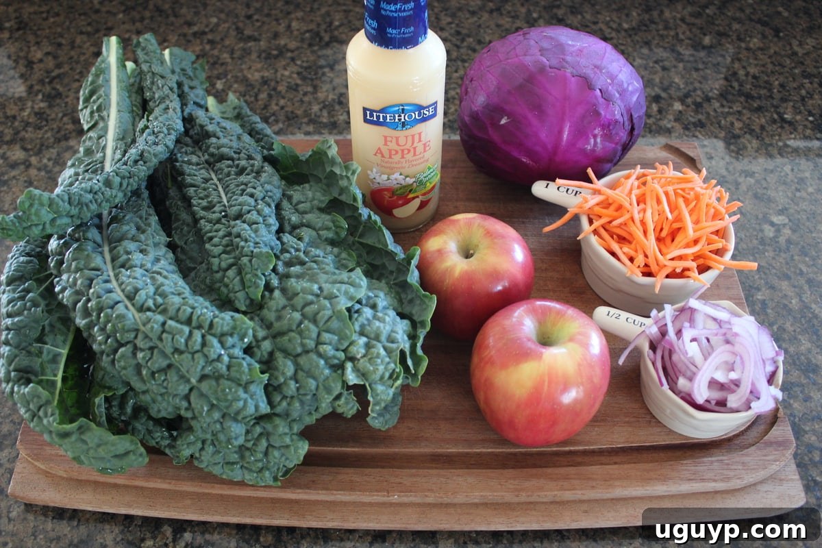 A vibrant selection of Kale Apple Slaw ingredients including kale, shredded red cabbage, thinly sliced red onion, grated Fuji apple, and julienned carrots, neatly arranged on a wooden board.