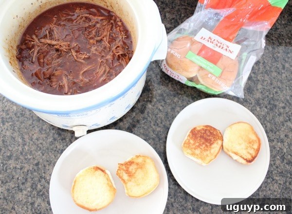 shredded brisket in sauce in a crock pot next to two plates with toasted hamburger buns