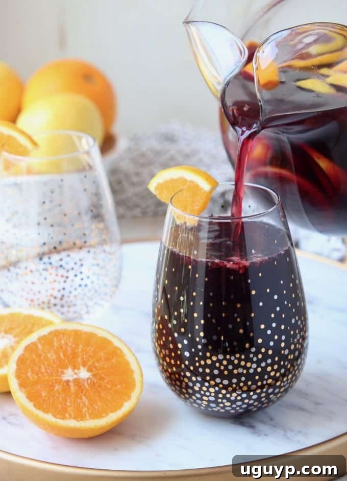 A glass of red sangria being poured from a pitcher, with oranges and ice visible.