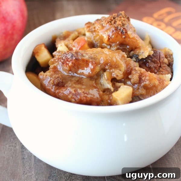 A close-up of baked caramel apple bread pudding in a white serving bowl, drizzled with caramel sauce and ready to be enjoyed.